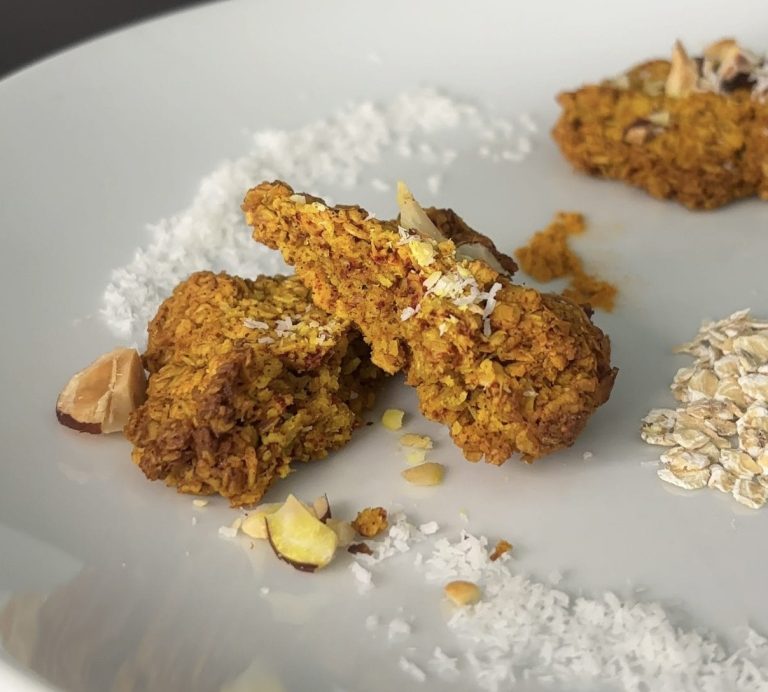 Gros plan sur un cookie healthy au curcuma, noix de coco et flocons d’avoine, cassé en deux pour révéler sa texture moelleuse, disposé sur une assiette blanche avec éclats d’amandes, coco râpée, curcuma et flocons d’avoine en décoration.