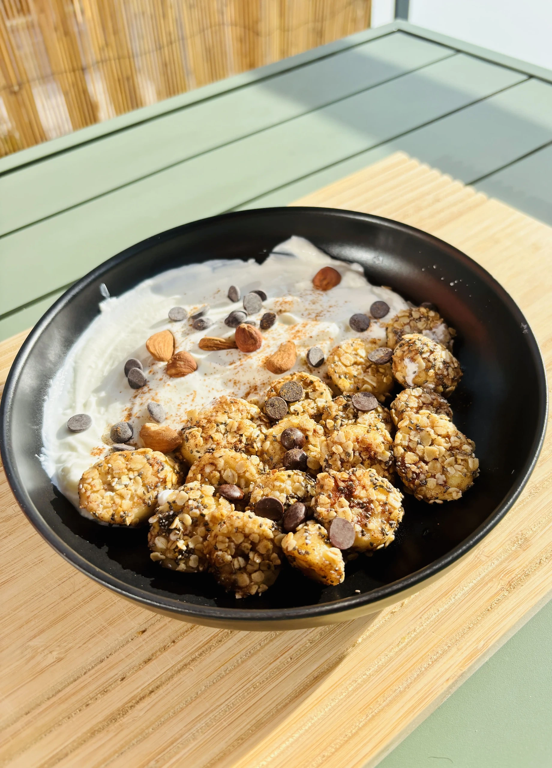 Bol noir contenant du skyr nature crémeux garni de banana oat balls dorées, saupoudrées de graines de chia et de flocons d’avoine, décoré d’amandes entières et de pépites de chocolat noir, posé sur une table en bois en plein soleil.
