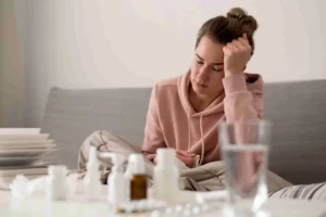 Femme assise sur un canapé, la tête appuyée sur la main, entourée de médicaments, illustrant la fatigue, le stress et un possible dérèglement du cortisol.
